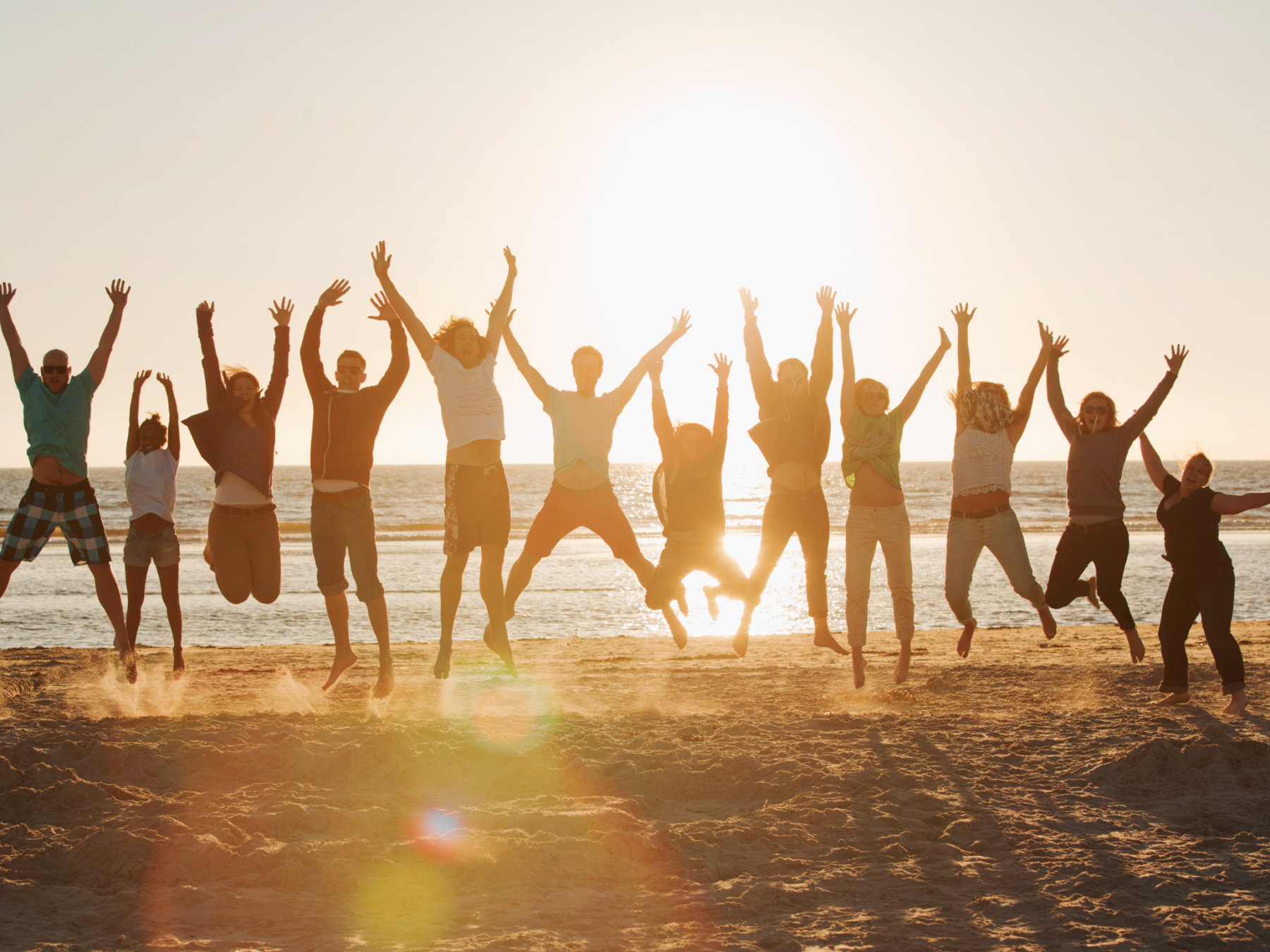 Picture of a group of people jumping in the air on a beach in front of the ocean. The sun is setting in the background